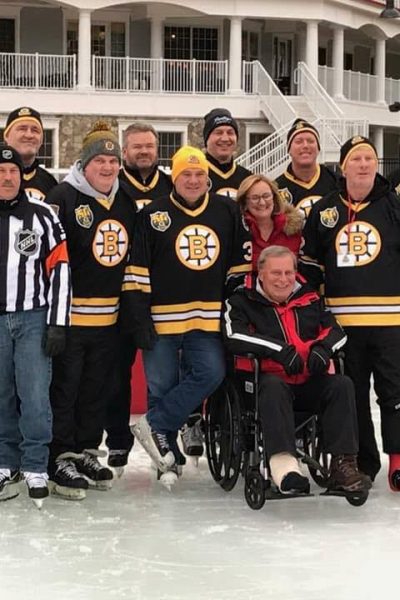 Nine individuals stand smiling (seven of whom are wearing Bruins jersey’s and one wearing a referee uniform) with a man sitting in a wheelchair with an ace wrap around his ankle in front of the group.