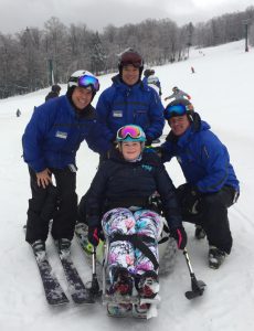 Three coaches wearing blue coats pose smiling behind an athlete sitting in bi-ski holding outriggers wearing colorful pants and goggles.