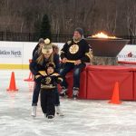 Individual skates with a toddler skating in front of her with a individual in a Bruins jersey sitting in the background on the barrier to a ceremonial fire pit.
