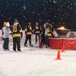 A group of individuals wearing Bruins jerseys stand on an ice rink around a ceremonial fire pit with snow falling