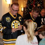 An individual wearing a Bruins jersey signs a small object using a sharpie for two children standing in front of them. Another individual, also wearing a Bruins jersey, is signing their autograph on something next to them.