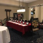 Silent auction table set up with a red tablecloth and ~15 item lists. Several items displayed around the table including two framed sports jerseys, a bicycle, and a large framed photograph of a football tackle.