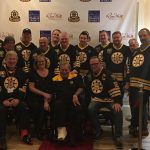 Group of 15 individuals wearing Bruins jerseys pose smiling with an older individual sitting in a wheelchair with a cast on right foot.