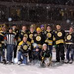 A group of individuals wearing Bruins jerseys and a referee stand/kneel smiling for the camera on an ice rink. Two children in sled hockey sleds pose in front of the individuals. It is evening and snowing.