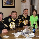Four individuals sitting around a dinner table wearing Bruins jerseys pose with a child standing between them wearing a green shirt.