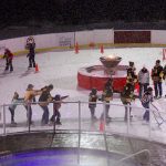 People skate in conga line around the ice rink in the evening while it is snowing.