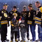 Five individuals wearing Bruins jerseys pose around two children on an ice rink. One child is wearing a hockey helmet and the other child is holding their helmet looking up at one of the individuals.
