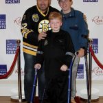 An individual wearing a Bruins jersey poses with two individuals standing with crutches on a red carpet.
