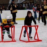 Two kids skate side-by-side using red walkers. One child is wearing a hockey helmet and Bruins outfit, and the other child is wearing a black hoodie and jeans. Three individuals watch smiling in the background.