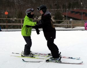 Two individuals on skis are facing each other with the coach’s skis straddling the athlete's skis. Their arms are interlocked in front of them and the coach is guiding the athlete down a slope.