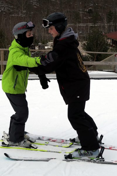 PAC coach skis backwards with an athlete in a bright yellow jacket skiing forward with skis parallel in between PAC member’s skis. Both individuals have their arms outstretched towards each other.