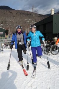 Two individuals are smiling standing on one ski and each is holding two outriggers. Each of the individuals has a left leg amputation.