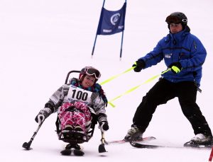 A smiling individual is seated in a bi-ski, skiing downhill with outriggers held out to each side, while a coach is skiing behind holding tethers.