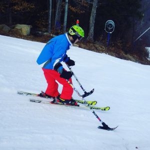 An individual is skiing downhill while standing on two skis and utilizing one outrigger in each hand.