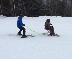 Individual riding ski bike down mountain with coach holding tethers from behind.