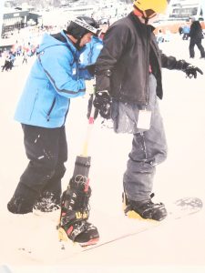  Two individuals are facing the same direction with the coach’s hands on the athlete’s back. The athlete is riding a snowboard with one leg strapped in and o an outrigger held by the athlete’s right arm strapped into the binding.