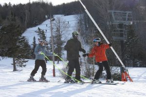 Three individuals skiing down the mountain. One coach from behind is holding tethers attached to the front of the athlete’s skis. Another coach is looking at the athlete while pointing down the mountain. The athlete is standing upright and looking forward. 