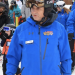 A close up view of a coach in a blue ski jacket, their face turned to the side, and goggles pulled up onto their helmet.