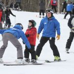 A child in a red jacket skis with one coach in front, skiing backwards with their skis straddling the child’s skis, and one coach in back skiing in wedge.