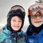 A child and an adult ski coach smile for a picture, each with their goggles pulled up on to their helmets.