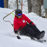 An individual in a red ski jacket skis down the slope while seated in a bi-ski, grasping outriggers.