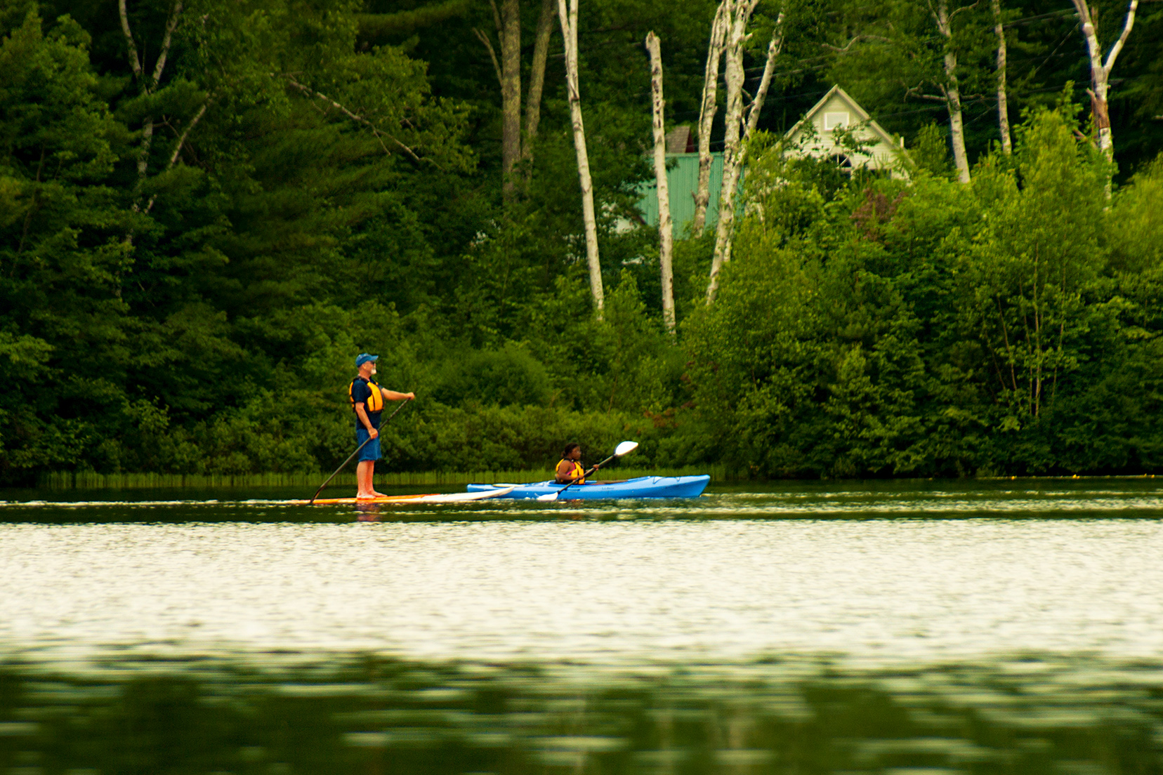 An individual is standing on a stand up paddle board while gripping a paddle. Just ahead, another individual is seated in a blue kayak gripping a paddle. Each individual is wearing a yellow life vest.