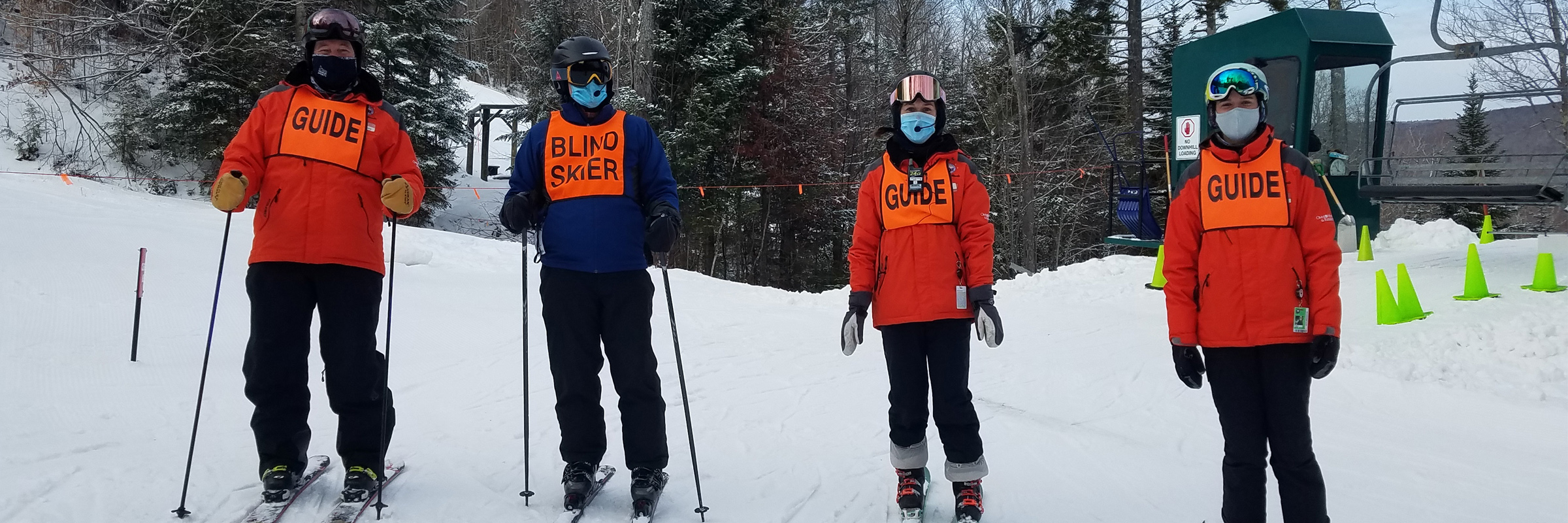 In this picture are 3 NEDS instructors all wearing orange jackets with an orange guide bib and a blind student wearing a blue jacket and an orange blind skier bib. This was during COVID, so each person is wearing a mask.